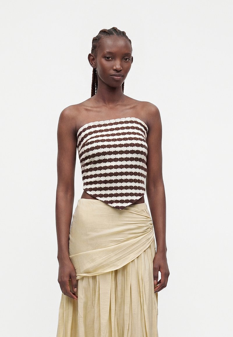 Femme portant un haut rayé marron et blanc sans manches et une jupe portefeuille beige plissée, debout devant un fond uni.