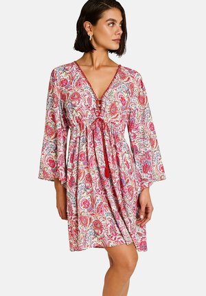 Femme portant une robe mi-longue à manches longues avec un motif paisley rouge et un décolleté en V profond noué par un cordon rouge.