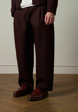 Bordeauxfarbene, maßgeschneiderte Hose mit Falten, aus glattem Stoff. Dazu dunkelbraune Leder-Loafer mit abgerundetem Zehenbereich und Riemen.