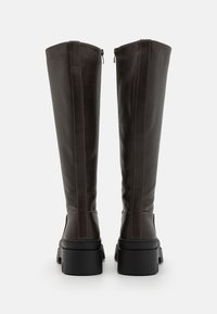 Bottes en cuir marron jusqu'aux genoux avec une texture lisse, présentant une fermeture éclair sur le côté et un large talon noir en bloc. Détails de couture visibles.
