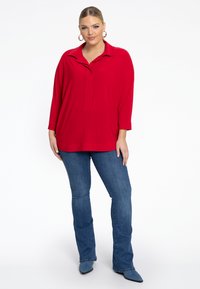 Roter übergroßer Pullover mit Kragen und Knopfleiste. Kombiniert mit hellblauen Schlagjeans und spitzen Schuhen. Glatte Textur.