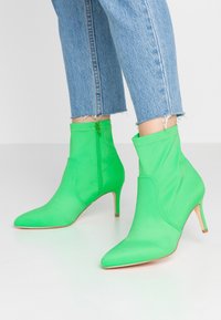 Zelené textilní kotníkové boty s špičatou špičkou a 6 cm jehlovým podpátkem, vybavené bočním zipem a hladkou texturou. Džíny s natrhnutým lemem.