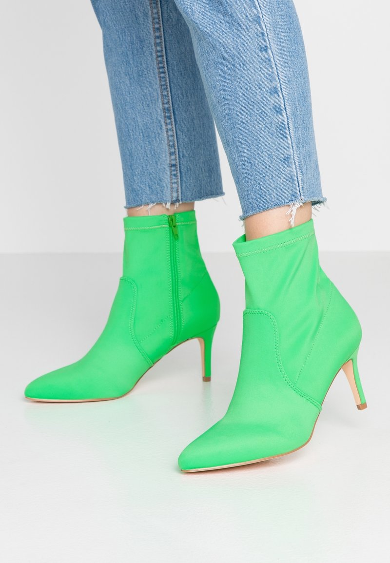 Zelené textilní kotníkové boty s špičatou špičkou a 6 cm jehlovým podpátkem, vybavené bočním zipem a hladkou texturou. Džíny s natrhnutým lemem.