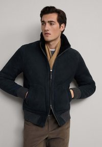 Tamno plavi zamak bomber jakna s krznom obloženim ovratnikom, rebrastim manžetama, patentnim zatvaračem i bočnim džepovima, uparen s bež maramom i sivim džemperom.