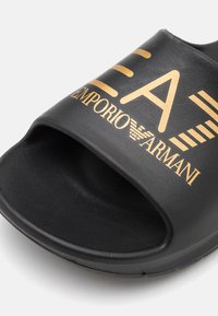 Black slide sandal made of textured synthetic material. Features gold embossed logo "EA7 Emporio Armani" across the strap.
