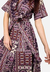 Žena v fialových květovaných zavinovacích šatech s krátkými rukávy, svázaných v pase, vidět ruce a střed těla.