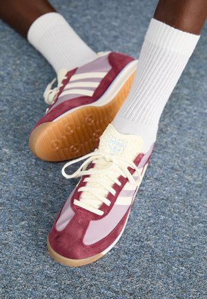 Personne portant des chaussettes blanches et des baskets Adidas bordeaux, lavande et crème avec semelles en gomme sur un sol moquetté bleu.