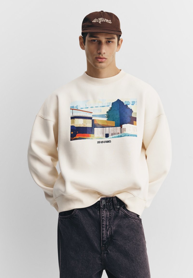Roomkleurige sweatshirt met een kleurrijke grafische print van abstracte gebouwen en bomen. Gecombineerd met donkergrijze spijkerbroek en een bruin petje.