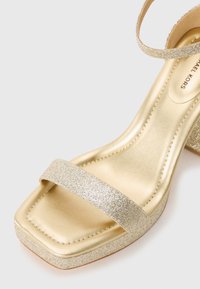 Zlata sandalna s peto v obliki bloka, z kvadratnim prstom, dvema bleščečima trakoma, gladkim kovinskim zaključkom in oblazinjeno podplatom.