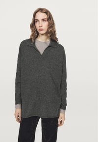 Giovane donna con capelli mossi che indossa un maglione largo grigio scuro sopra a una camicia grigio chiaro e pantaloni scuri, in piedi su uno sfondo chiaro e uniforme.