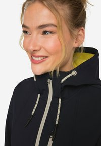 Veste de marine avec capuche, dotée de fermetures éclair et de cordons de serrage gris clair, accentuée par une doublure intérieure jaune. Texture de tissu douce visible.
