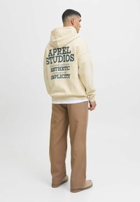 Cream hoodie with "APREL STUDIOS" logo on the back in dark green text. Worn with brown wide-leg pants and beige footwear.