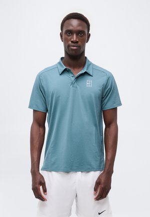 Homme portant un polo à manches courtes couleur sarcelle avec un petit logo blanc sur la poitrine gauche et un short blanc avec un Swoosh Nike noir.