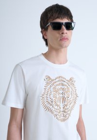 Camiseta blanca con un intrincado diseño de cortes en forma de cara de tigre de color marrón. Presenta mangas cortas y un cuello redondo. Gafas de sol negras con forma angular.