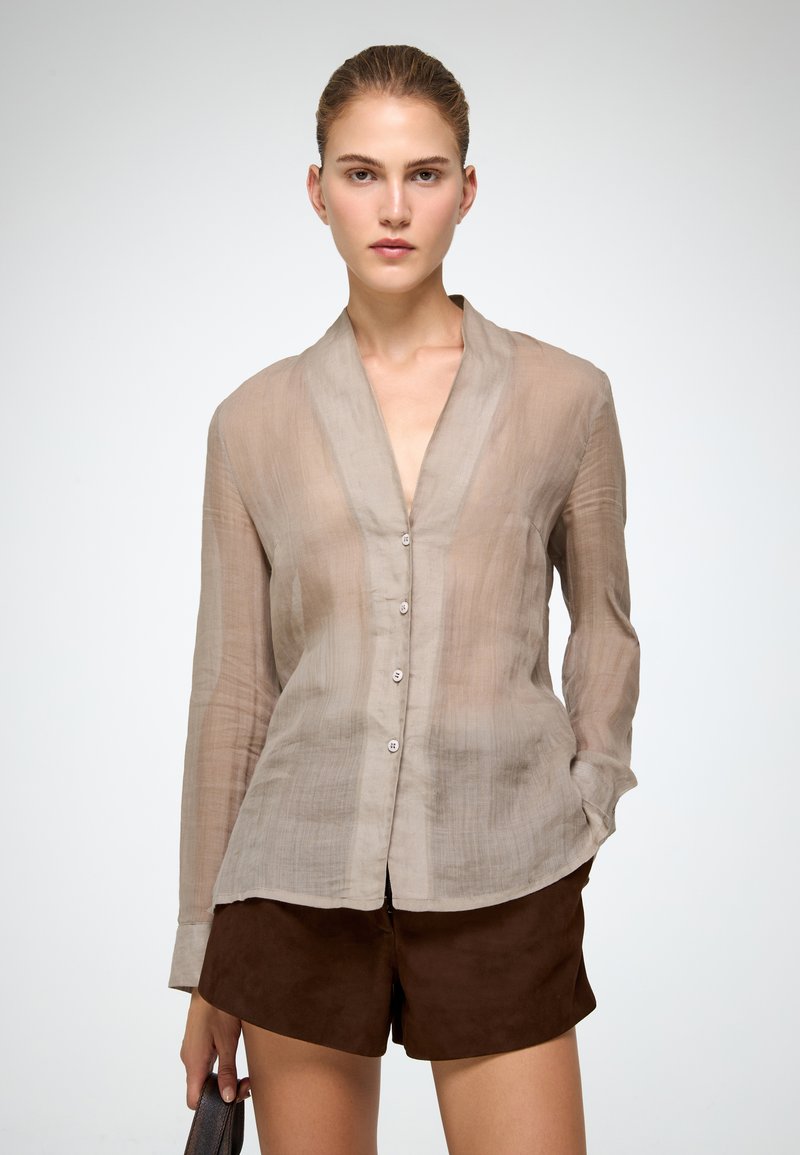 Camicia beige trasparente con bottoni, scollo a V e maniche lunghe; texture e trasparenza sottili, abbinata a pantaloni corti in suede marrone.