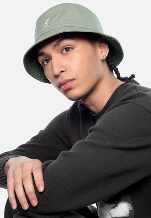 Jong persoon met gevlochten haar, een groene vissershoed, een zwarte trui en gouden creolen, met armen rustend en iets wegkijkend.