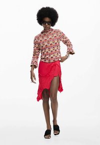 Langärmeliges Oberteil mit Muster in warmen Farbtönen, kombiniert mit einem roten Rock, der dekorative Blumenstickerei und einen seitlichen Schlitz aufweist. Schwarze Sandalen runden das Outfit ab.