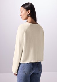 Maglione color crema con una finitura morbida e testurizzata, caratterizzato da un ampio scollo e maniche raglan, abbinato a jeans in denim azzurro chiaro.