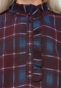 Közelkép egy bordó és kék kockás ingről, fodros díszítéssel a gombolópult mentén és magas gallérral, amit egy világos hajú személy visel.