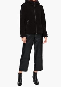 Giacca in pile nera con zip, cappuccio e tasche laterali; abbinata a pantaloni in pelle nera a vita alta e stivali con tacco spesso alla caviglia.