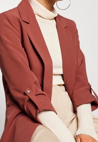 Rostfärgad blazer med långa ärmar och knäppningar vid ärmsluten, buren över en krämfärgad ribbad polokrage. Slät tyg med skräddarsydd design.