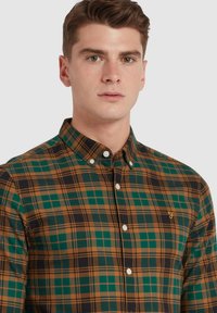 Groene en bruine geruite overhemd met button-down kraag, voorzien van witte knopen en een klein geel logo op de borst.