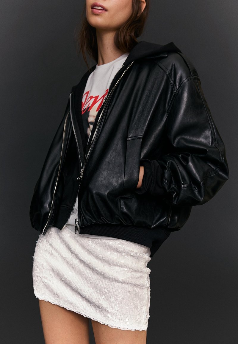 Femme portant une veste en cuir noire, un t-shirt graphique blanc et une mini-jupe blanche texturée, posant devant un fond sombre.