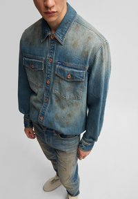 Denim košulja u izblijedjeloj plavoj boji, s dva džepa na prsima, smešnim dugmadima i blago iznošenim izgledom, uparena s usklađenim trapericama.