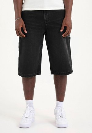 Uomo che indossa pantaloncini di jeans neri fino al ginocchio, calzini bianchi alla caviglia, sneakers bianche e un braccialetto d'argento al polso sinistro.