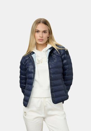 Giovane donna con lunghi capelli biondi che indossa una giacca imbottita blu navy sopra una felpa bianca con cappuccio e pantaloni della tuta bianchi su uno sfondo semplice.