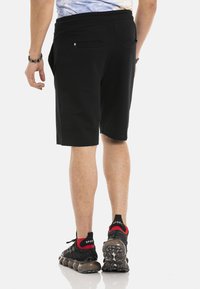 Zwarte knielange shorts met een gladde textuur, voorzien van twee achterzakken. Gedragen met zwart-rode sneakers met gestructureerde zolen.