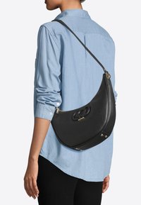 Sac à main en cuir noir avec un design incurvé, garnitures argentées et un logo bien en vue à l'avant. Il est doté d'une bandoulière longue et ajustable.