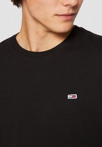 Musta puuvillapaita pyöreällä kaula-aukolla, jossa on pieni kirjailtu logo punaisena, valkoisena ja sinisenä vasemmassa rinnassa.