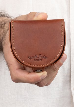 Brown leather coin wallet with curved shape, visible stitching, and embossed logo. Closed with a snap button, held in a hand.