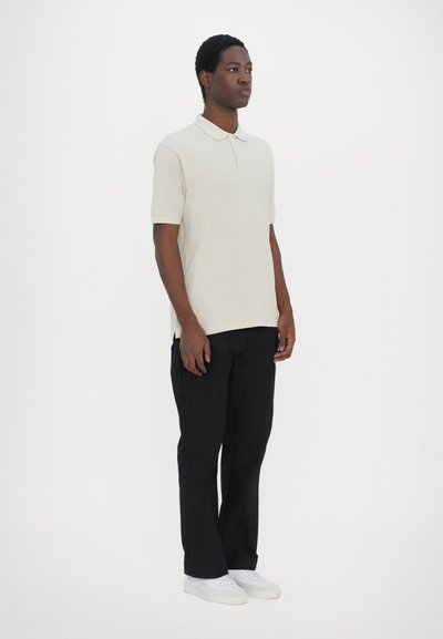 Polo shirt beige à manches courtes, associé à un pantalon noir et des baskets blanches. Design simple, coupe régulière et texture lisse.