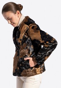Kobieta w brązowo-czarnym wzorzystym sztucznym futrze, nosząca beżowy golf, z rękami w kieszeniach kurtki, patrząca w dół.