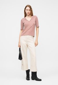 Γυναίκα με κυματιστά μαλλιά που φοράει πάνω μέρος μπλούζας με κόκκινες και άσπρες ρίγες σε σχήμα V, κρεμ παντελόνι με φαρδιά μπατζάκια και σχέδιο μανσετών, μαύρες μπότες και κρατάει μια μικρή μαύρη τσάντα.