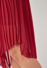 Falda roja plisada hecha de un tejido ligero y semitransparente. Presenta un diseño fluido con una textura suave y un dobladillo desigual.