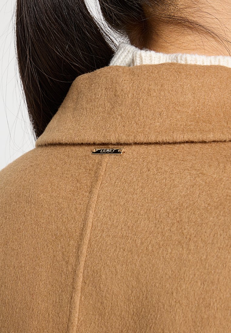 Brązowy wełniany płaszcz o gładkiej fakturze, z kołnierzem i małą złotą metką marki blisko dekoltu.