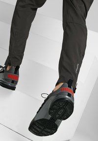 Un pantalon de sport gris présente une ouverture zippée à la cheville et un design épuré. Les chaussures sont noires et grises avec un logo "PUMA" rouge bien visible sur le talon.