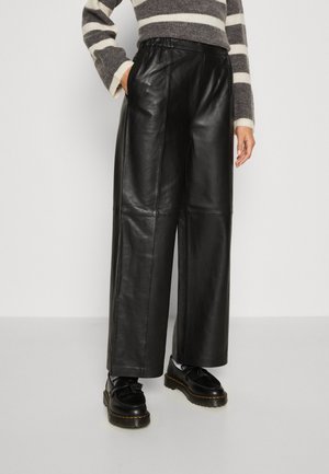 Person iført sorte læderbukser med vide ben, sorte loafers og en grå-hvidstribet sweater med hænderne afslappet.