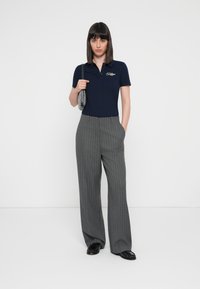Polo de color navy con mangas cortas y detalle del logotipo, combinado con pantalones de pierna ancha a rayas grises. La modelo sostiene un bolso de mano con un estampado.