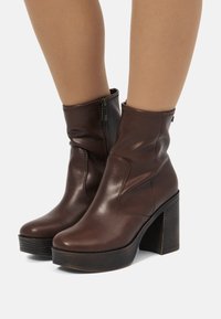 Stivali alla caviglia in pelle marrone con altezze raggruppate, chiusura laterale con zip e tacco spesso e irregolare. Texture liscia con un design elegante.