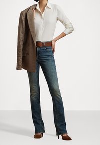 Kvinna iklädd en vit skjorta instoppad i blå jeans, håller en brun rutig kavaj över ena axeln, med ett brunt bälte och klackar.