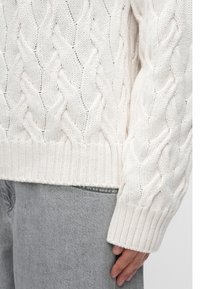 Personne portant un pull torsadé crème et un jean gris clair, visible de la taille jusqu’au milieu des cuisses, une main reposant sur le côté.