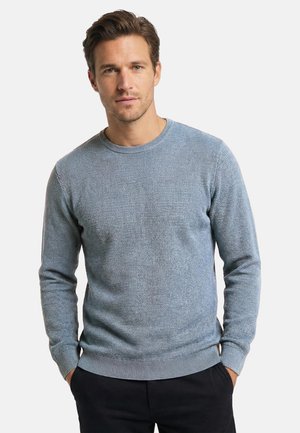 Uomo con capelli corti castani che indossa un maglione azzurro chiaro traforato e pantaloni neri, in piedi con le mani in tasca davanti a uno sfondo semplice.