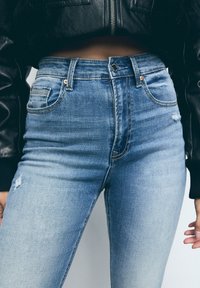 Jeans de mezclilla azul de cintura alta con un ligero estiramiento, que presentan bolsillos frontales, remaches de cobre y un desgaste mínimo en el muslo.