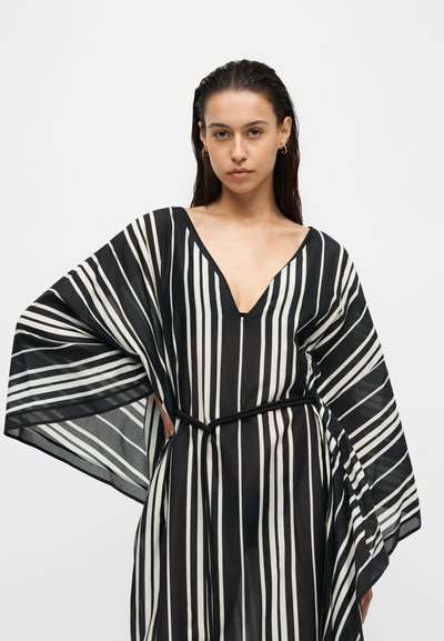 Femme aux cheveux foncés porte une robe rayée verticalement noire et blanche avec de larges manches et une ceinture noire, debout devant un fond uni.