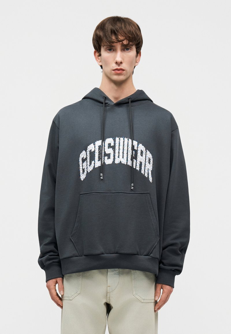 Young man wearing a dark gray GCDSWEAR hoodie with front pocket and beige pants, standing against a plain light background.