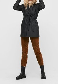 Svart, bälte, långärmad kappa med knappstängning; bärs över bruna kordfloretbyxor och svarta ankelstövlar. Slät textur, avslappnad design.